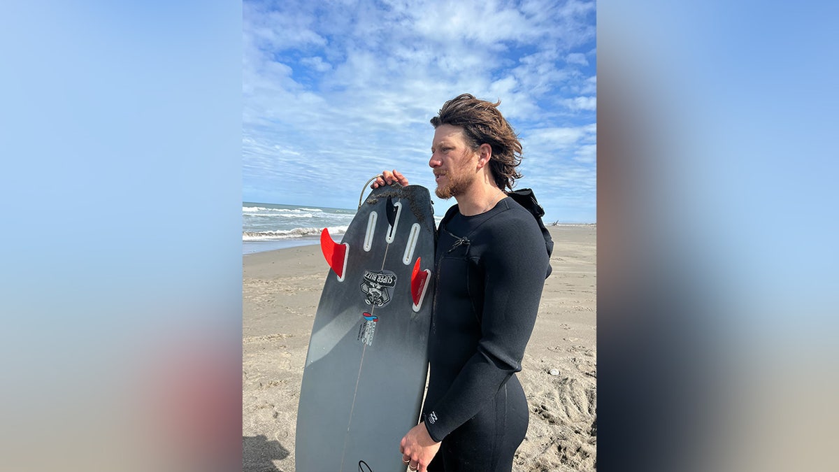 Jared Trainor stands on the beach with a surfboard