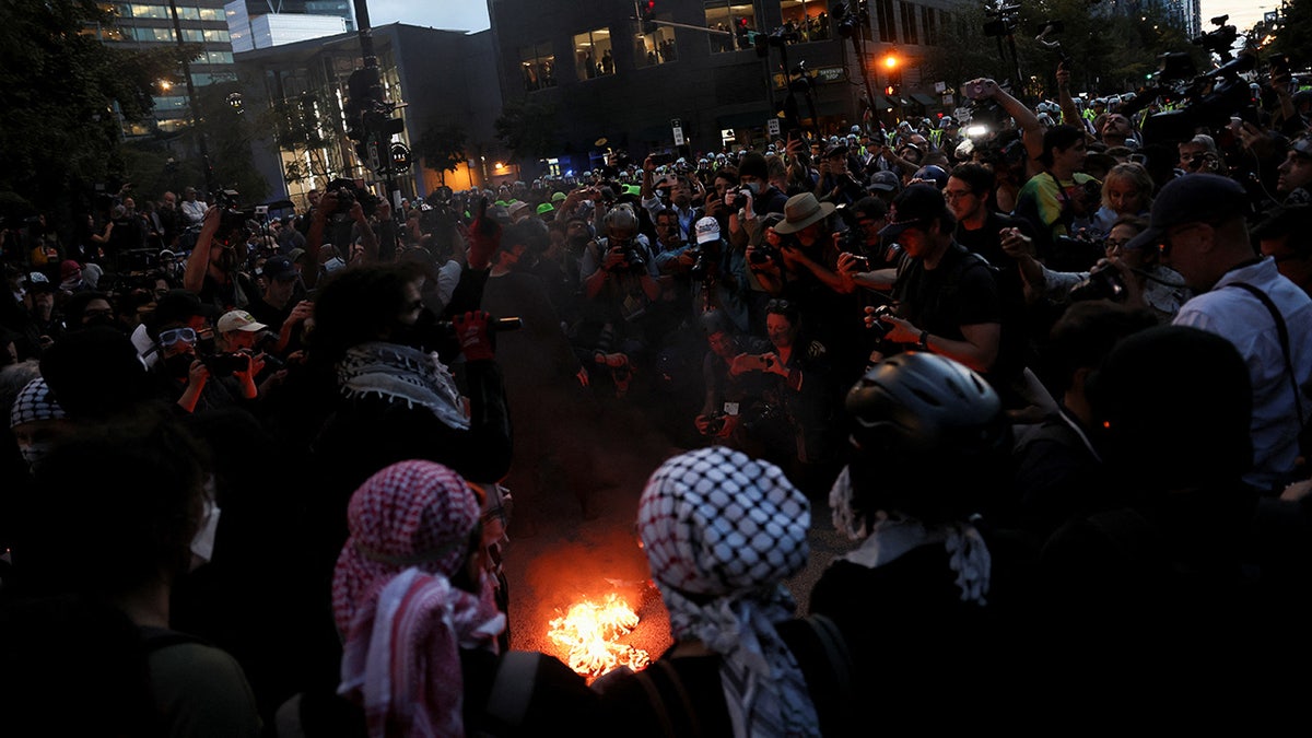 Members of the media shoot as protestors light a flag on fire on the sidelines of the Democratic National Convention