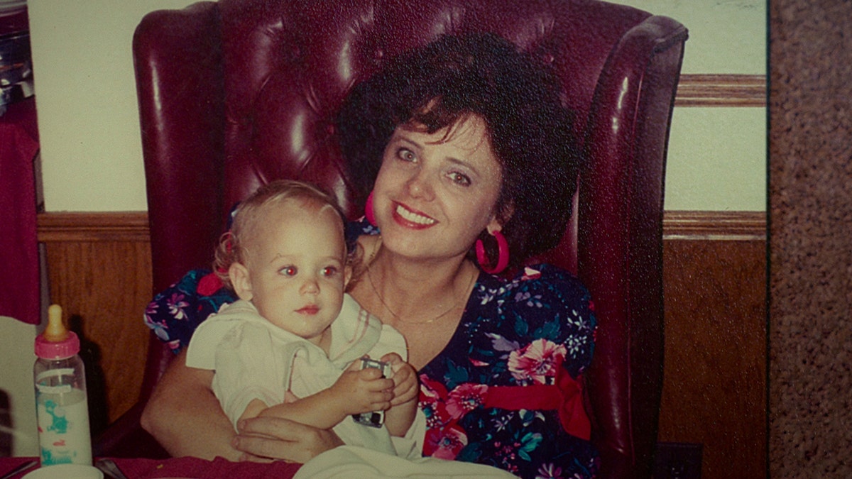 Patsy Ramsey holding a baby JonBenét Ramsey wearing a floral dress and smiling.