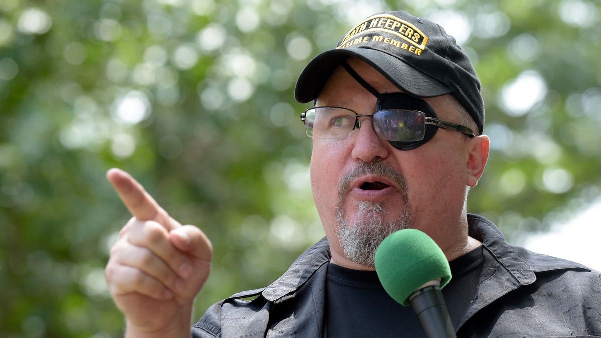 Stewart Rhodes wearing an eyepatch, holding a mic, and pointing his finger while giving a speech