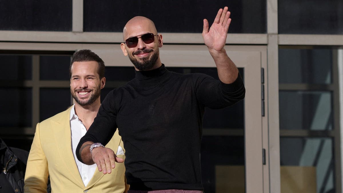 Andrew tate, wearing a black tutle neck and sunglasses, waves after leaving a Romanian courthosue, with his brother Tristan walking behind him in a tan sportscoat